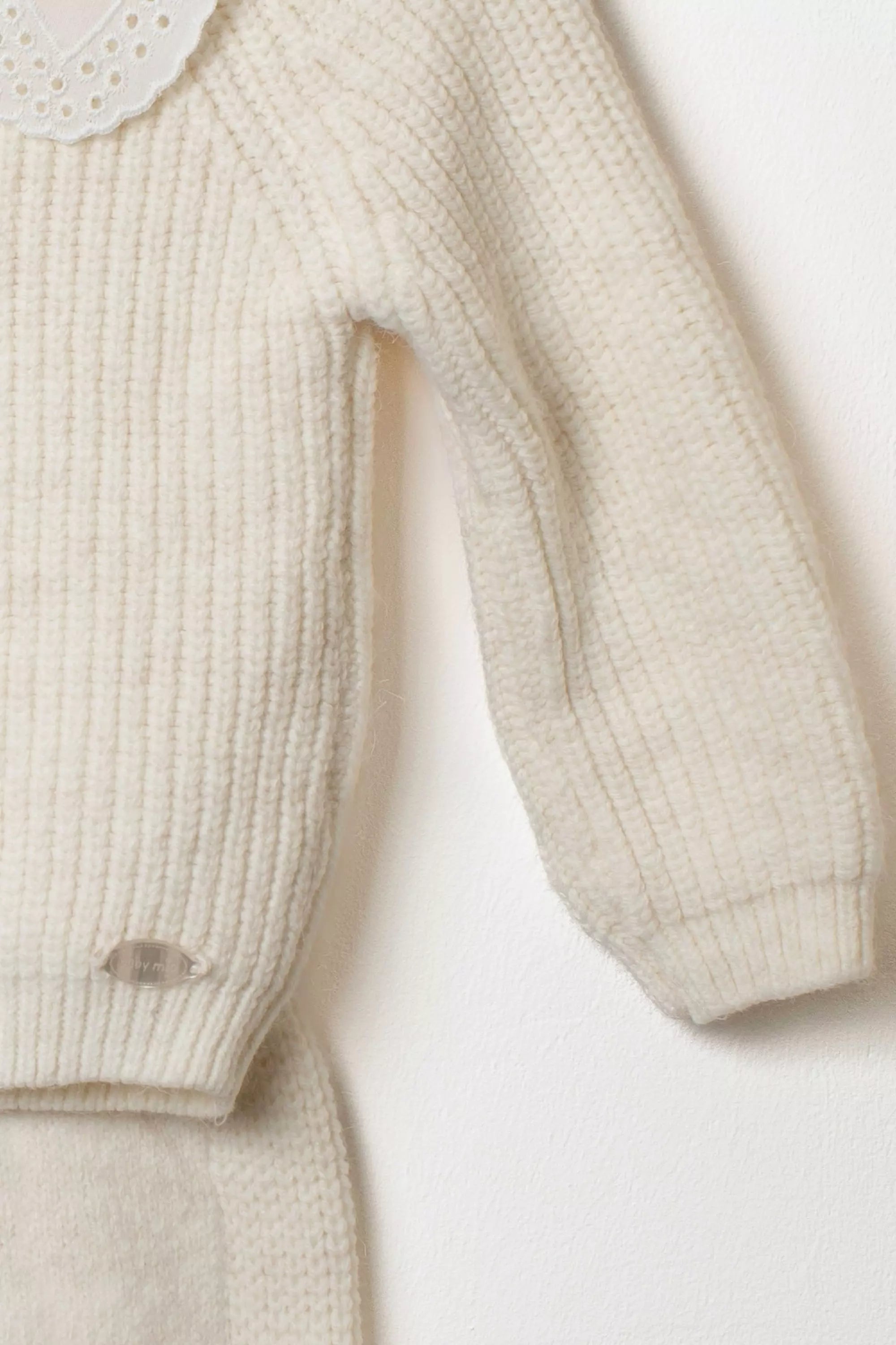 Nahaufnahme des Ärmels des cremefarbenen Strickpullovers mit Rippstrickmuster und kleinem Logo Detail