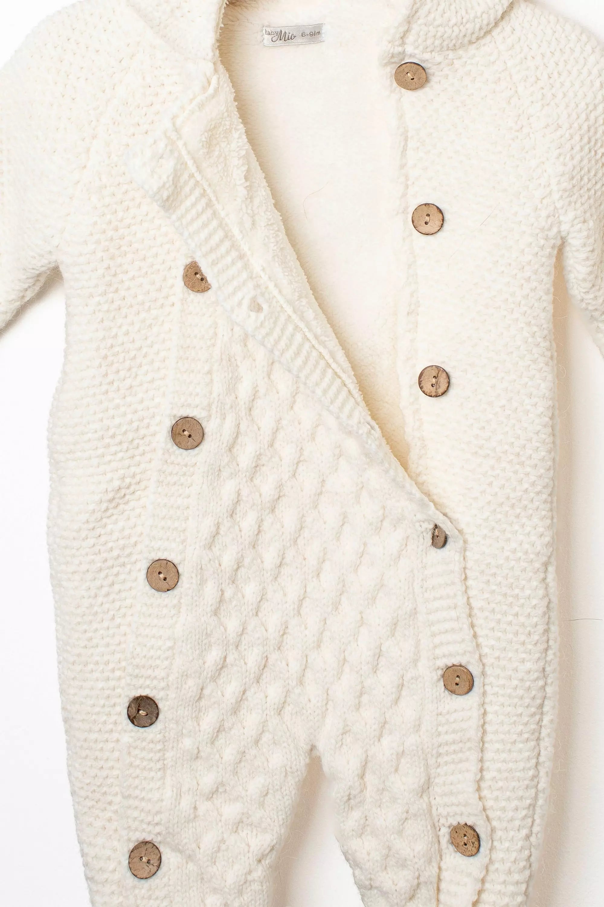 Nahaufnahme des geöffneten cremefarbenen Baby Strick-Overalls mit Zopfmuster, weichem Teddyfutter und rustikalen Holzknöpfen.