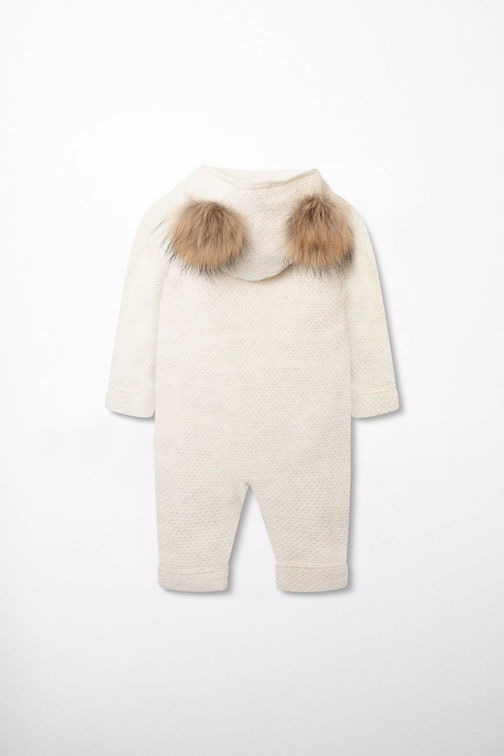 Rückansicht des cremefarbenen Baby Winter-Overalls aus Strick mit niedlicher Kapuze mit zwei Webpelz-Bommeln.