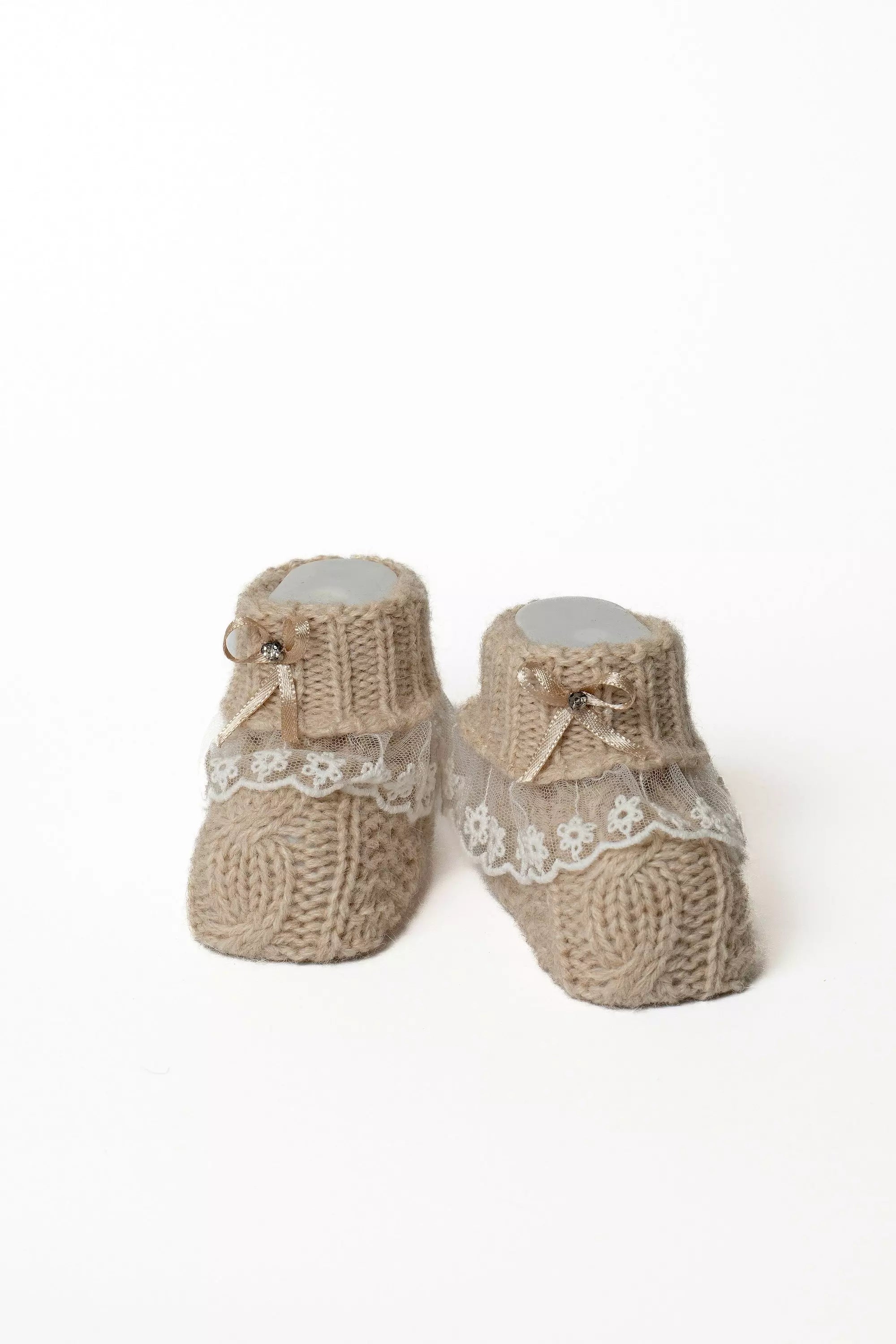 Paar braune Babystrickschuhe mit feiner weißer Spitze, eleganter Satinschleife und kleinem Glitzerdetail.