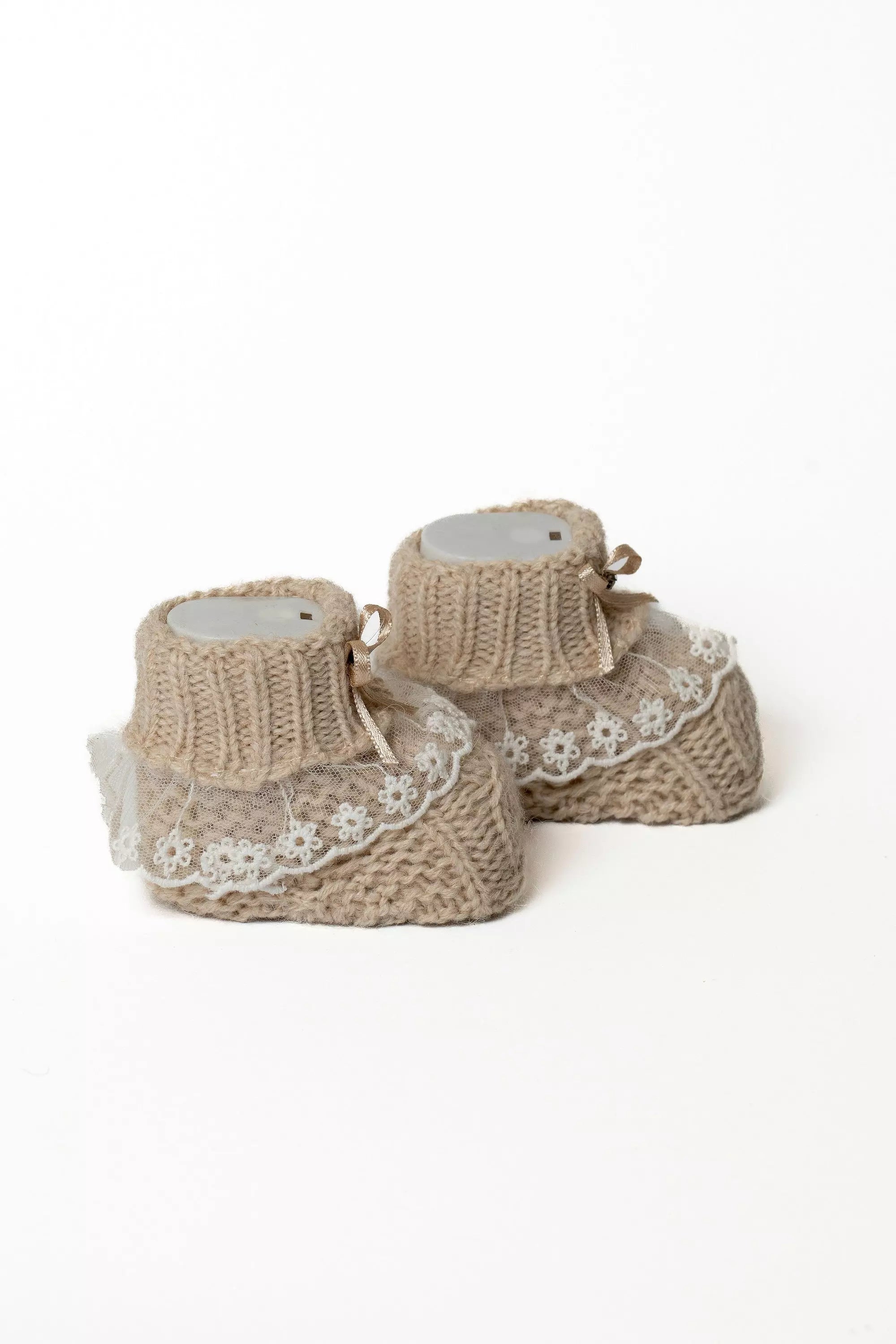 Paar beigefarbene Babystrickschuhe mit eleganter Spitze und kleiner Schleife, auf weißem Hintergrund.