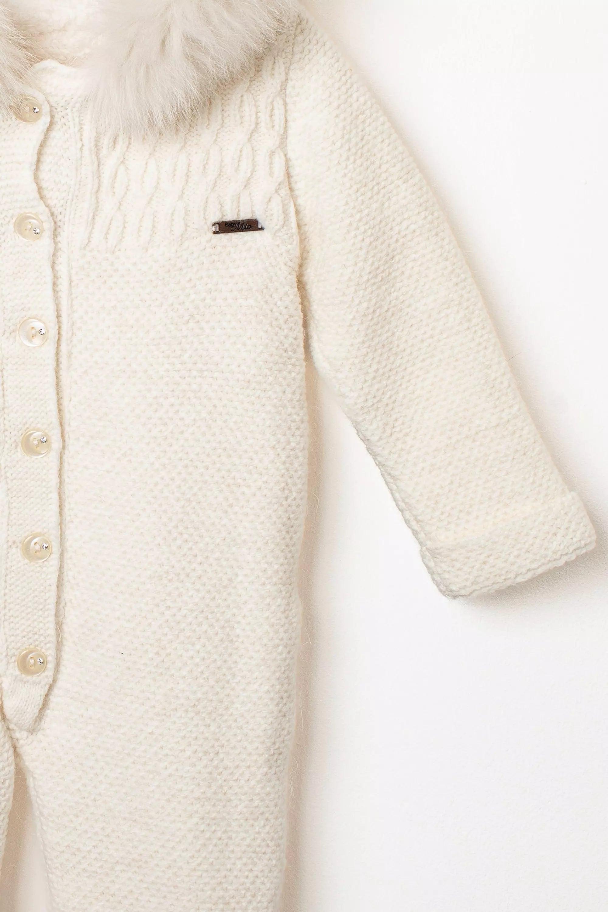 Nahaufnahme des Ärmels und der Brustpartie des weißen Strick-Overalls mit Zopfmuster und Logo-Emblem