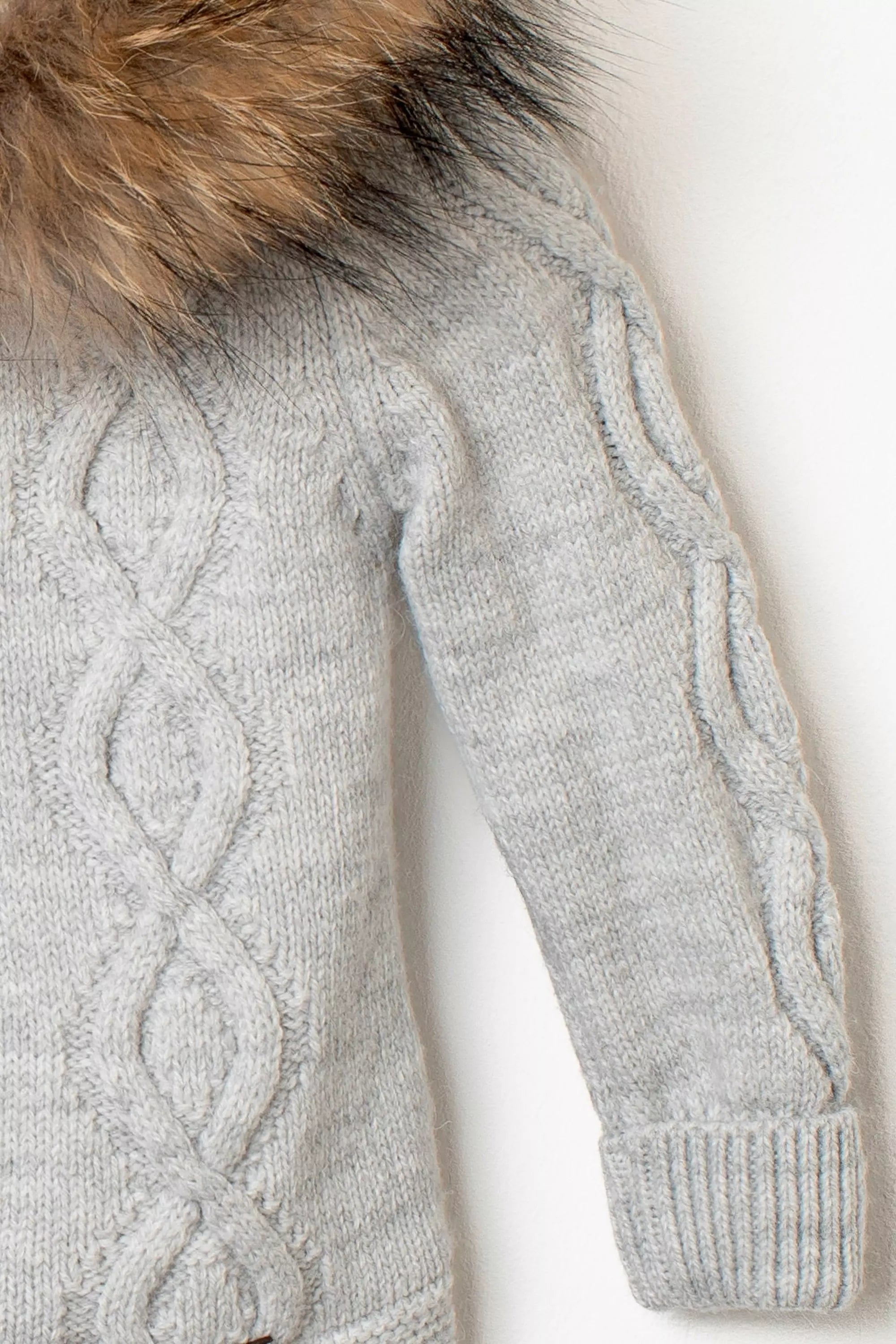 Nahaufnahme des Ärmels der grauen Baby-Strickjacke, mit detailreichem Zopfmuster und Rippbündchen, sowie Pelzbesatz an der Kapuze