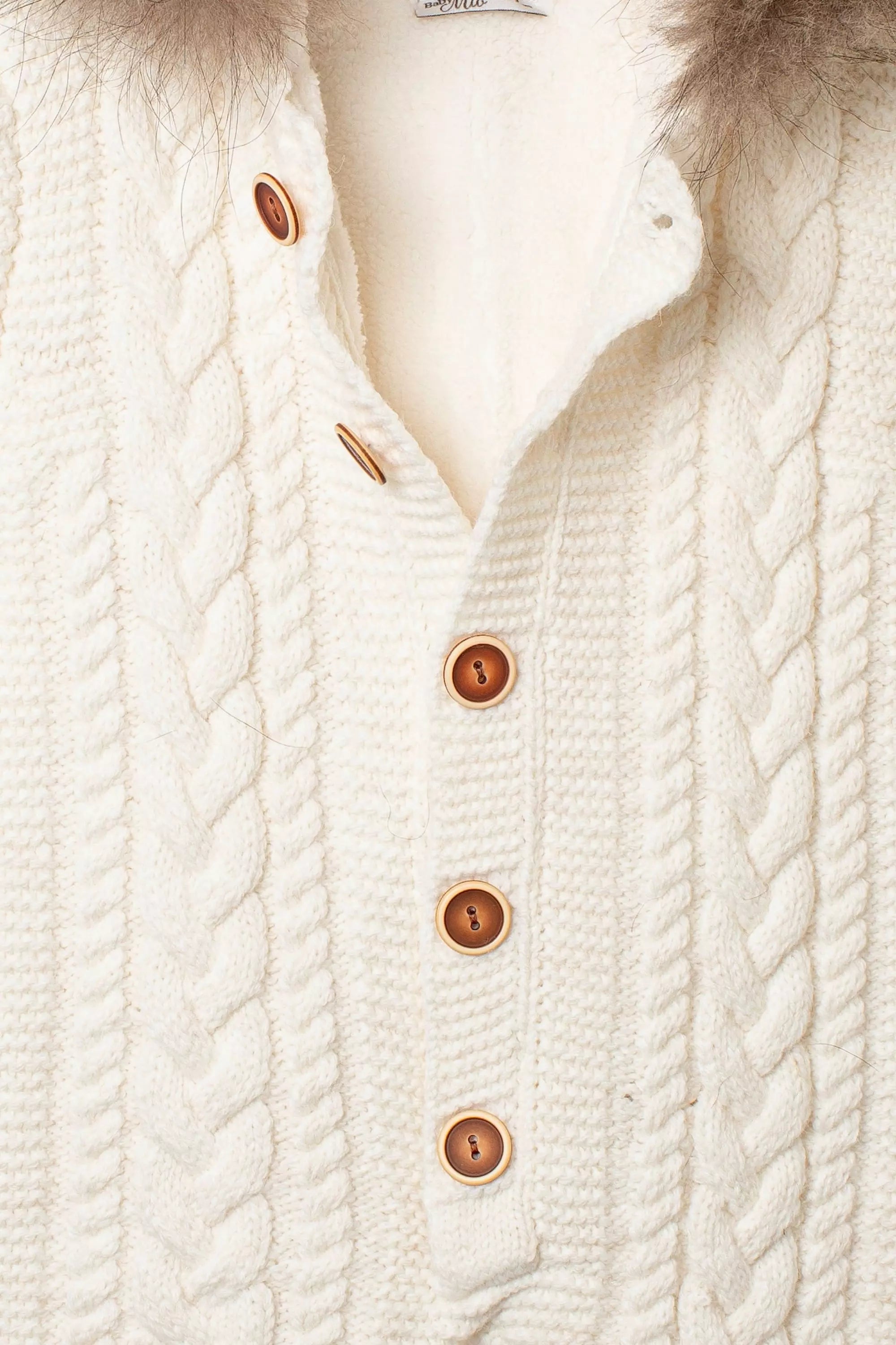Nahaufnahme der Knopfleiste des creme-weißen Baby-Overalls, mit Zopfmuster und braunen Holzknöpfen auf flauschigem Innenfutter