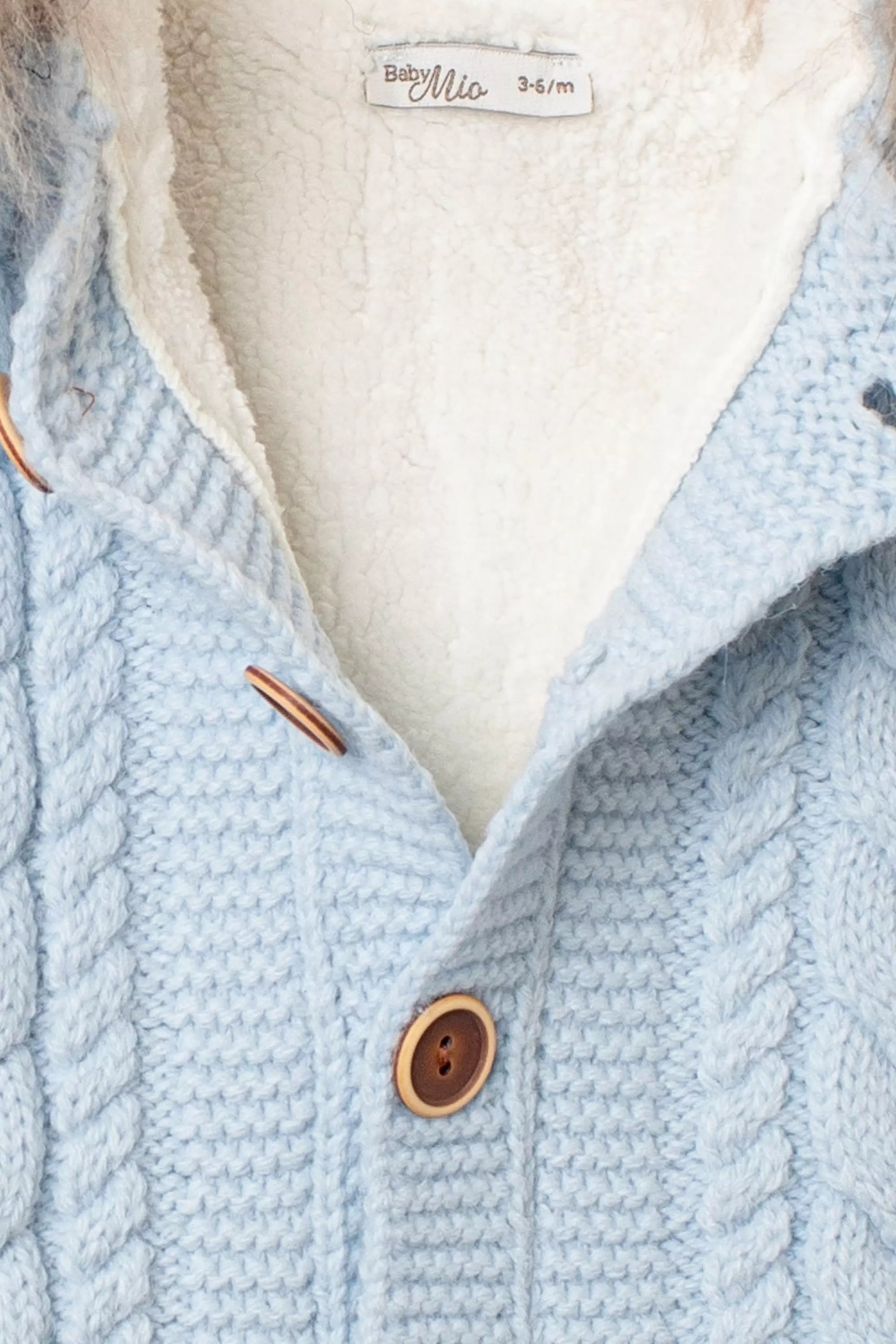 Nahaufnahme der Knopfleiste des hellblauen Baby-Overalls, mit Zopfmuster, Holzknopf und hellem, flauschigem Innenfutter