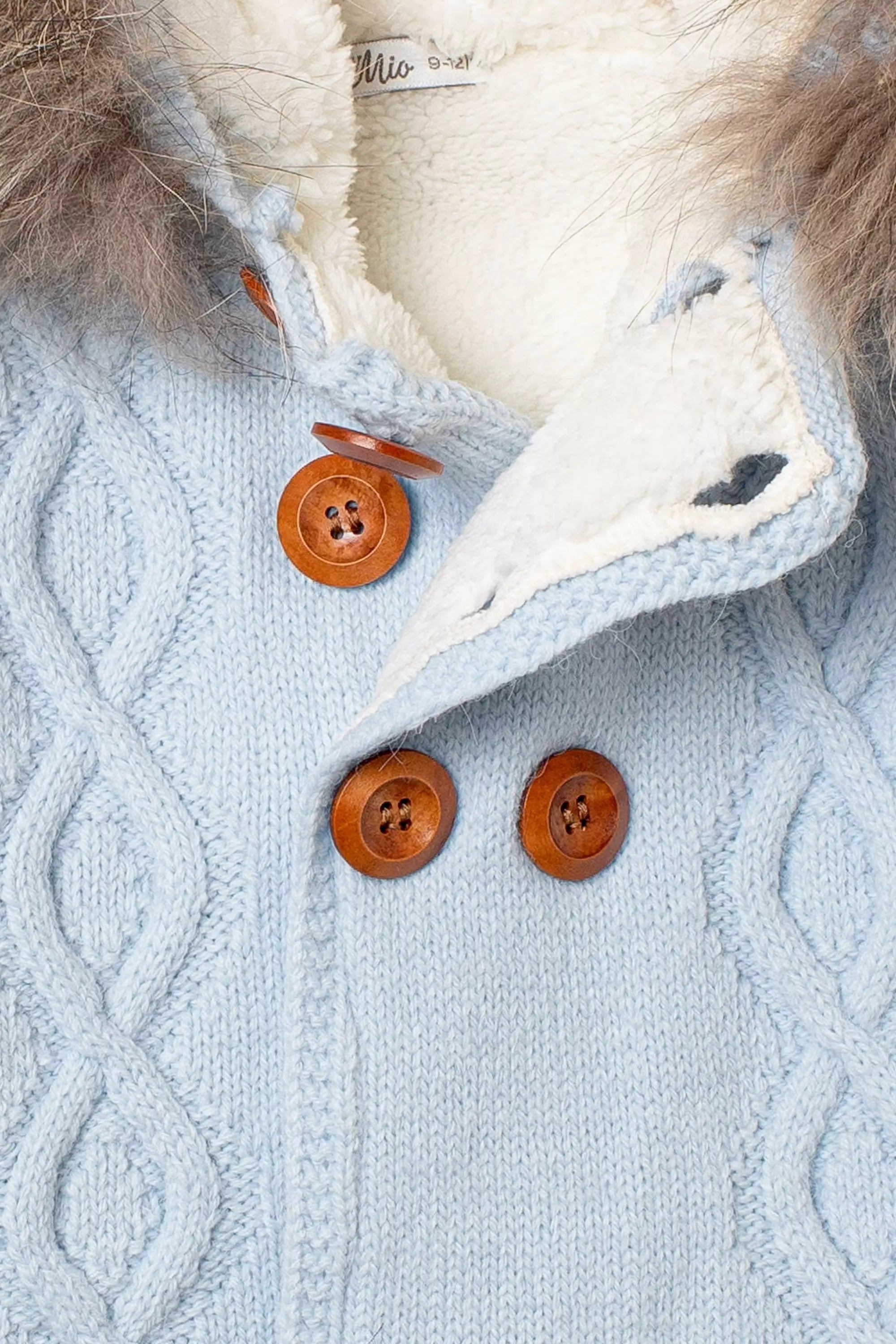 Nahaufnahme der Knopfleiste der hellblauen Baby-Strickjacke, mit Zopfmuster, rustikalen Holzknöpfen und flauschigem weißem Innenfutter