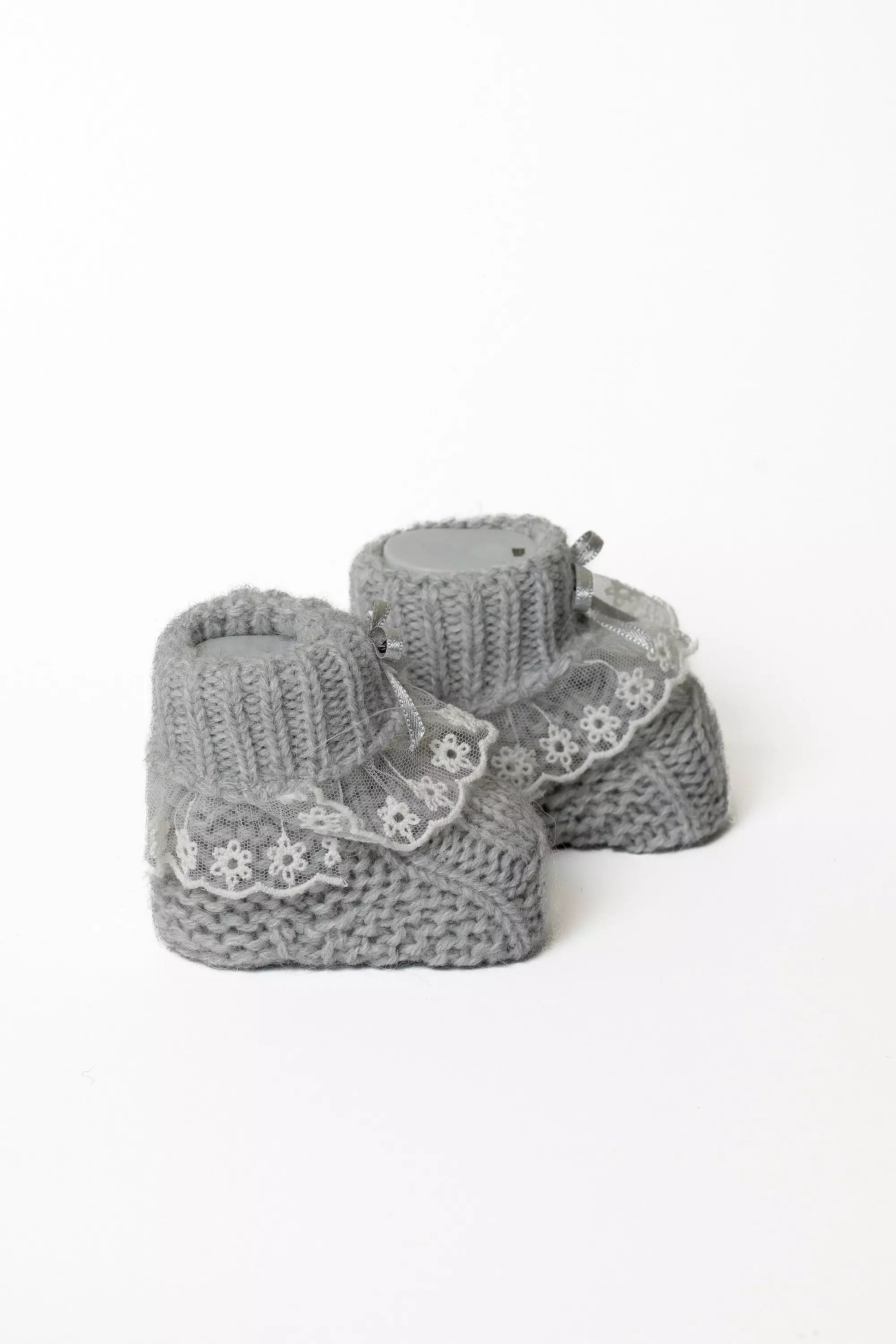 Paar graue Babystrickschuhe mit weißer Spitze und silberner Schleife, ideal für Mädchen-Outfits.