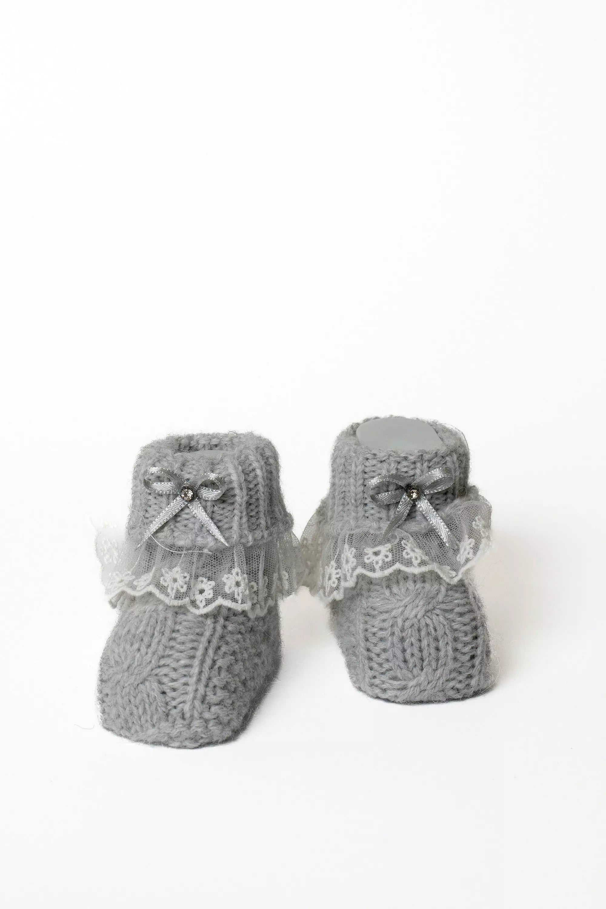 Paar graue Babystrickschuhe mit Zopfmuster, weißer Spitze und eleganter silberner Schleife für Neugeborene.