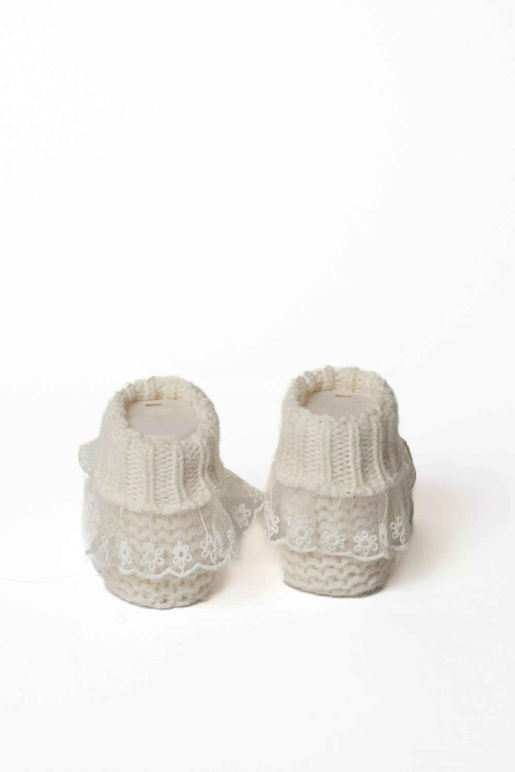 Paar cremeweiße Babystrickschuhe von hinten, mit Rippbündchen und großer weißer Spitzenverzierung.