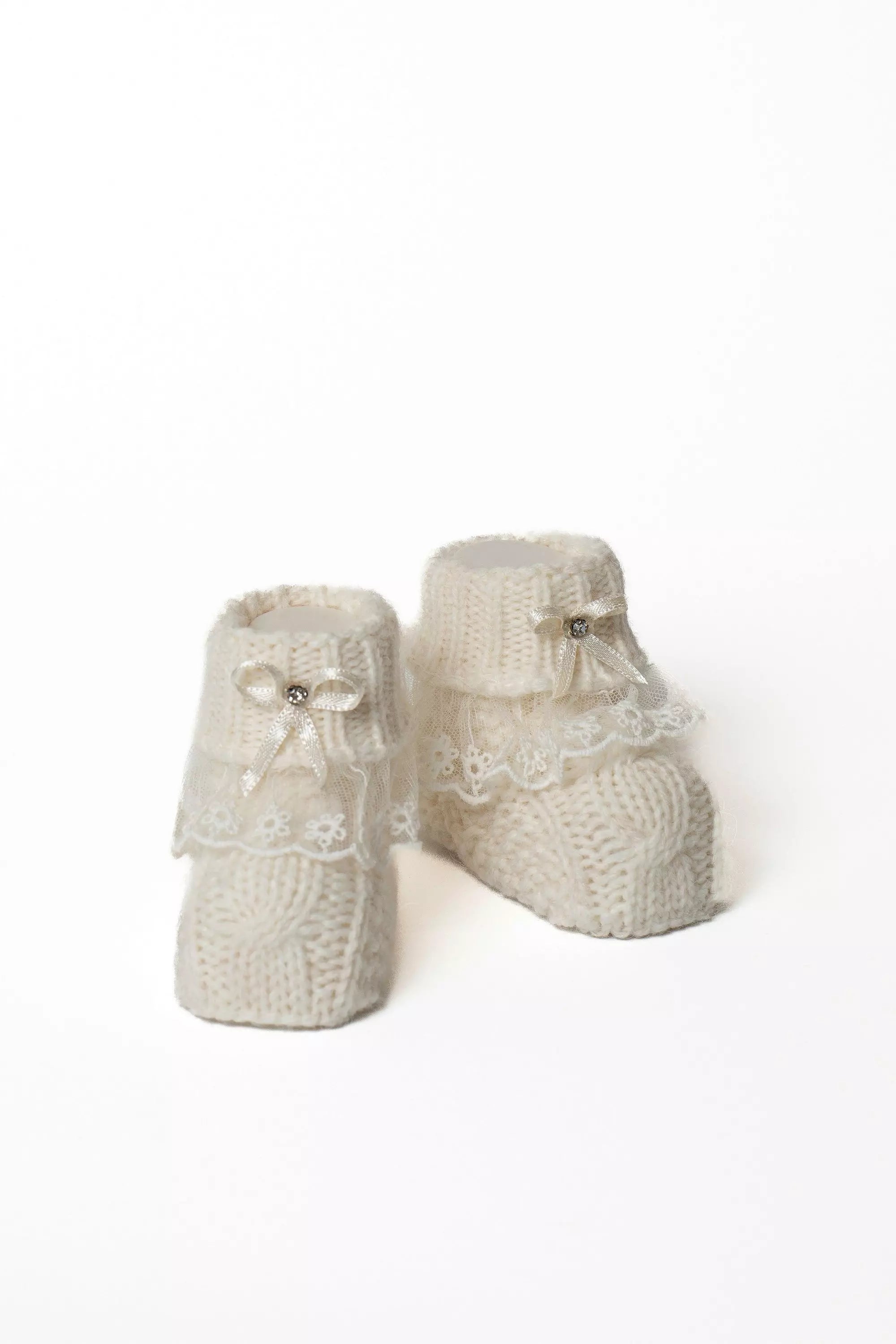 Paar weiße Babystrickschuhe mit Zopfmuster, dekorativer weißer Spitze und kleiner Satinschleife mit Glitzerstein.